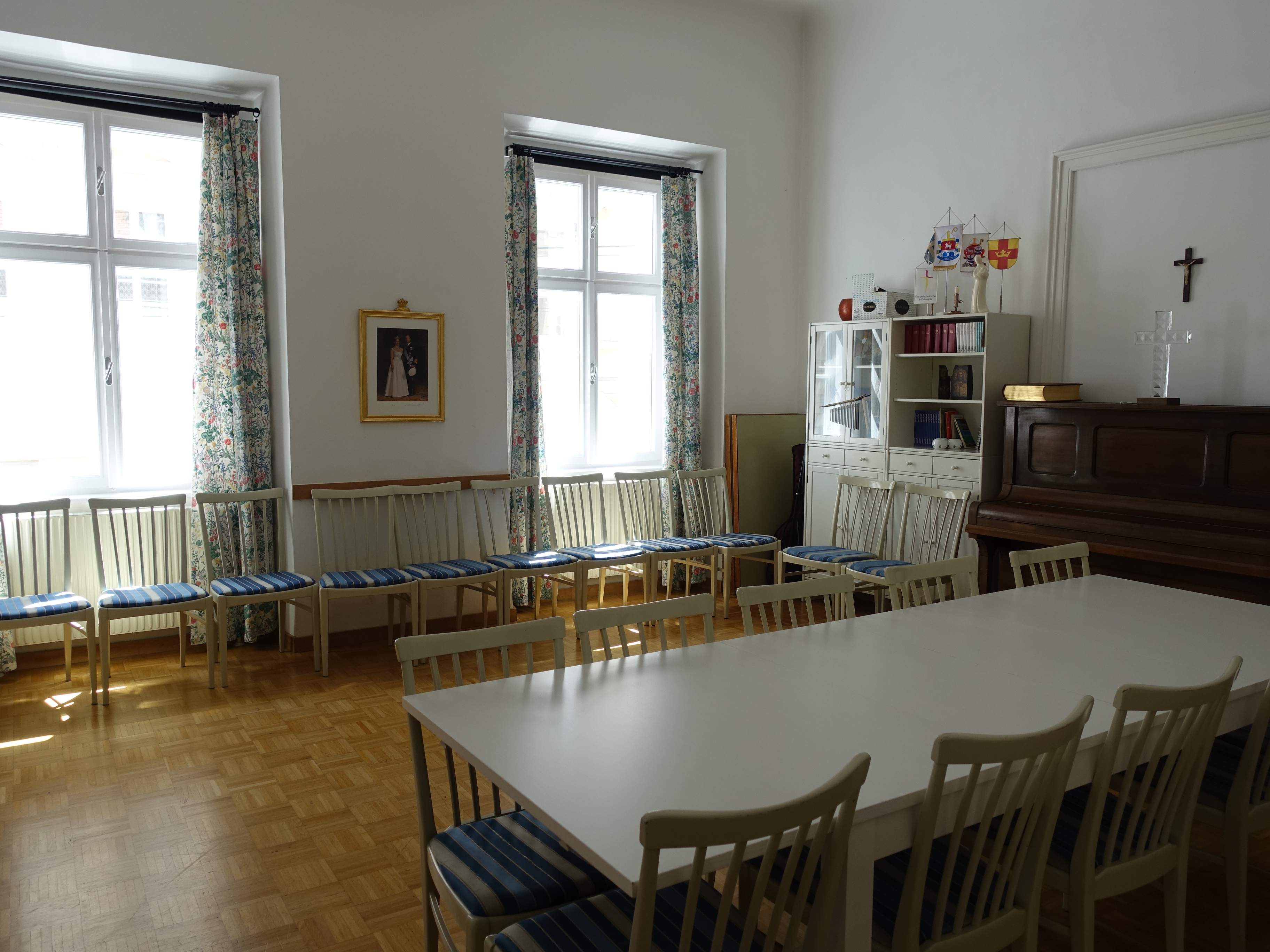 Kuva salista, jossa näkyy pöytiä ja tuoleja sekä piano, taustalla ikkunat.